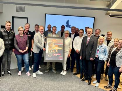 23 Personen stehen in einem großen Besprechungsraum. In der Mitte stehen ein Mann und eine Frau, die ein Plakat zum Dreckburg Open Air 2026 in der Hand halten